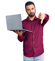 Young hispanic man holding laptop with angry face, negative sign showing dislike with thumbs down,...