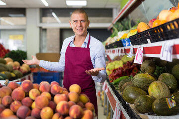 Male worker in apron standing in salesroom of greengrocer and looking in camera.