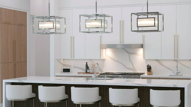 Interior of luxurious white furnished modern kitchen with expansive marble countertop and five chairs around. Three silver led chandeliers of square shape hanging on ceiling