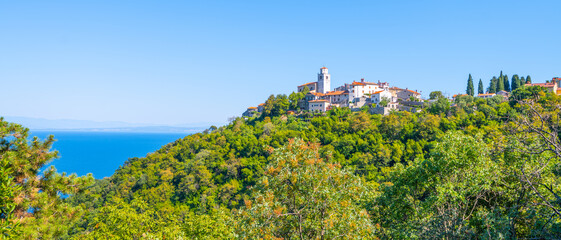 Naklejka premium Lush greenery surrounds a charming village perched on a hill overlooking the Adriatic Sea along the Istria Peninsula. Bright weather enhances the picturesque coastal landscape.
