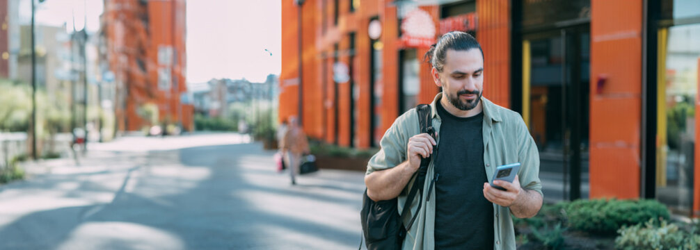A young Caucasian man with a phone in his hands on a bright city street.