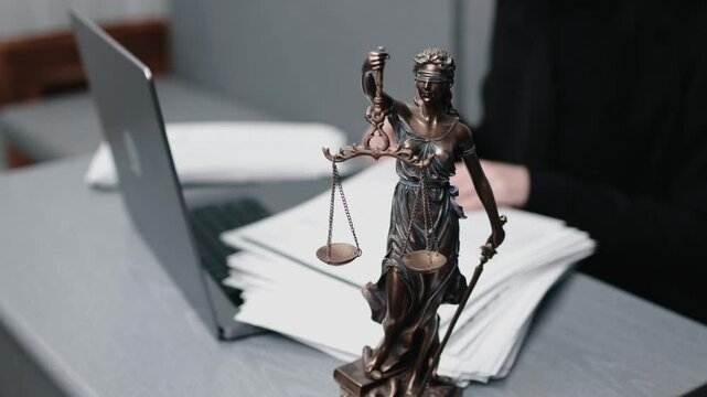 A lawyer works at their desk, sorting papers and entering data on a laptop. The statue of Themis stands nearby. Concept of legal decisions, work, and court proceedings.