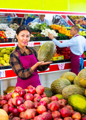 Asian woman in uniform standing in greengrocer and holding melon in hands. Her male colleague working in background.