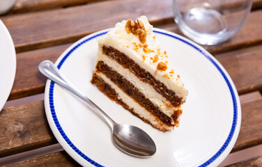 There is tea treat on plate. Piece of carrot cake made with addition of aromatic spices cinnamon and ginger. Layers of pie alternate with sour cream, and pastry is decorated with walnuts on top.