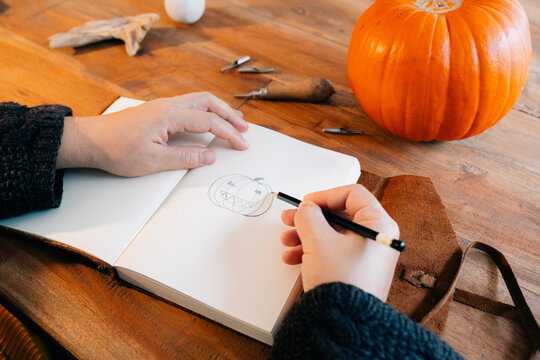 Person drawing a jack-o'-lantern sketch with a pumpkin on a table