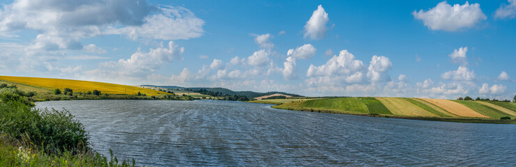 A panoramic view across the reservoir to hilly fields with rapeseed, sunflower, and lush cereal crops under a bright blue sky with clouds.