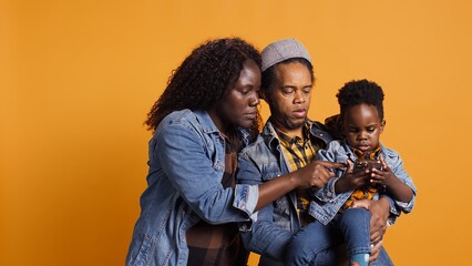 Cheerful african american family taking pictures against yellow background, posing with their small child. Smiling mother and father takes photos on smartphone, making memories. Camera B.