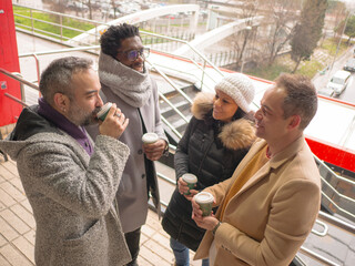 Four office workers taking a coffee break in a lively business district. The group consists of one Black male, a woman, and two Hispanic individuals. They are looking at each other and engaging in