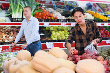 Focused adult asian woman visiting greengrocery store, choosing sweet pomegranates on counter..