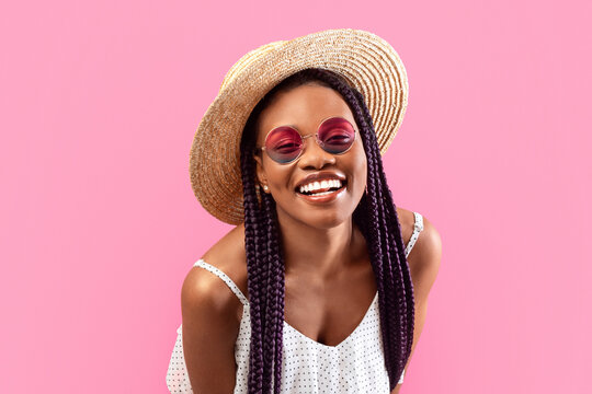Portrait of beautiful black lady with braids wearing trendy summer outfit, straw hat and sunglasses, smiling on pink studio background. Pretty African American woman enjoying summertime