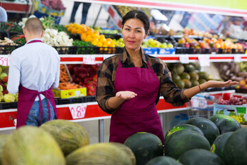 Smiling Asian saleswoman inviting to fruit and vegetable store, offering fresh organic produce..
