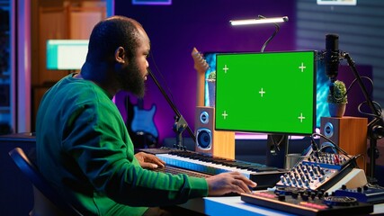 Songwriter operates on synthesizer piano keys next to isolated display, composing new music with instruments and stereo tools in home studio. Artist producer works with green screen. Camera B.