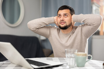 Annoyed unhappy bearded guy working at home at table with laptop, covering ears with his hands...