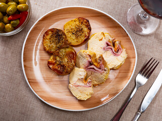 There is chicken meat on plate, cooked with bacon filling and garnished with pieces of fried potatoes. Next to main course there is snack bowl with pickled green olives
