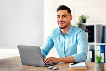 Fototapeta premium Attractive young Arab businessman using laptop pc at his desk in home office. Cheerful Eastern guy entrepreneur typing on computer keyboard, having in online conference at workplace