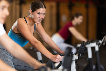 Fototapeta premium Girl hardly performs exercises on exercise bicycle during fitness class. Gym and fitness equipment, visitors are engaged in physical exercises during training