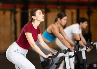 Fototapeta premium Young woman in athletic uniform during a group workout on an exercise bike in the gym. European woman on a cardio machine