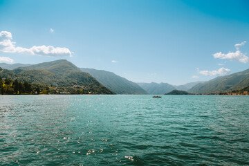Breathtaking panoramic views of Lake Como feature crystal clear waters reflecting sunlight, with a lone motorboat gliding across the surface against a majestic mountain backdrop