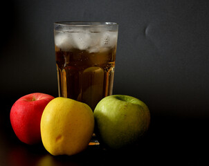 Apple juice from a mixture of fruits of different varieties in a tall faceted glass with ice on a black background.