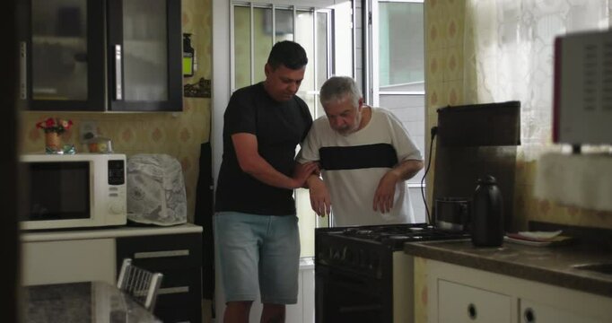 A man supporting an elderly man with dementia or age-related illness walking forward in a kitchen, showing care, assistance, and compassion in a home environment