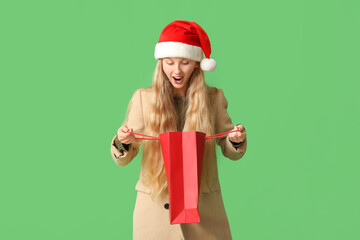Beautiful young shocked woman in warm winter clothes and Santa hat with shopping bag on green background