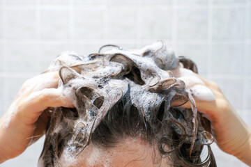 Naklejka premium A young woman washes her hair with shampoo on gray tiles background, front view.