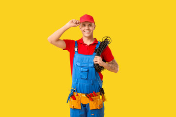 Young male electrician with tools and cable on yellow background