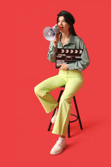 Young shocked female film director with movie clapper and megaphone screaming on chair against red...