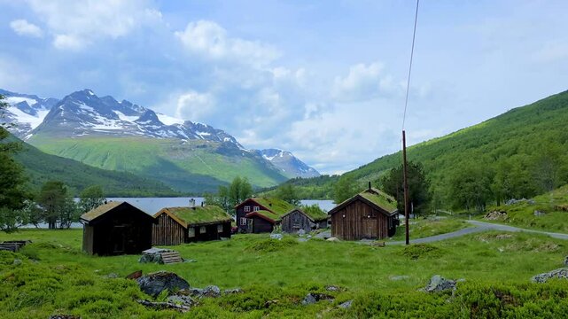 Experience the serene beauty of Norways lush greenery, nestled beside a tranquil lake. Quaint wooden cabins harmonize with majestic mountains under a clear blue sky, Innerdalen