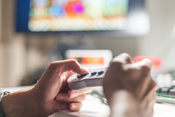 Close-up of hands holding a retro gaming controller with a blurred TV screen in the background. Perfect for gaming, nostalgia, and entertainment-related projects.