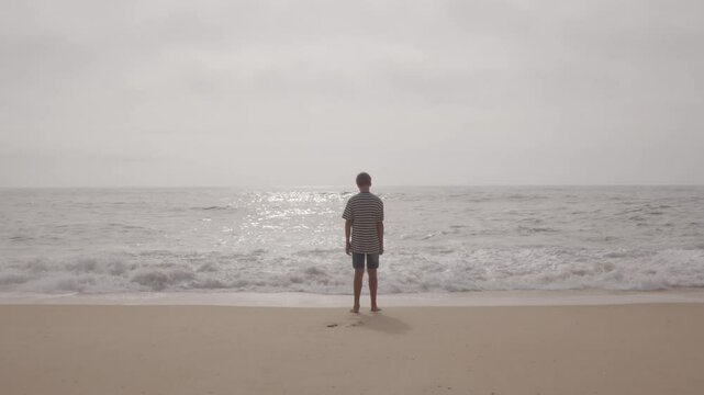 Teenage male standing on sandy shore, gazing pensively across overcast seascape with distant ocean waves under cloudy sky. Spain, Galicia