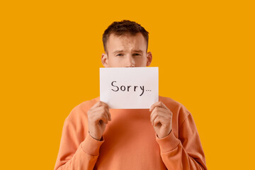 Young man holding paper with word SORRY on orange background