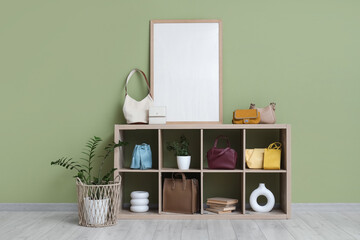 Shelf unit with stylish bags, blank frame and plants near green wall in room