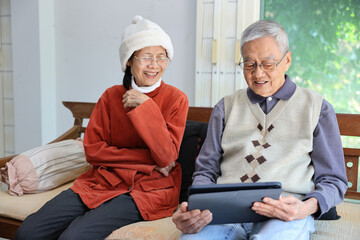 Senior asian couple are laughing and sitting on the sofa while watching funny stand up comedy show from streaming digital tablet during winter time for mental health and happiness in marriage