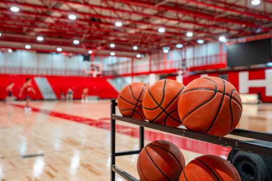 Close up photo of basketballs on storage rack inside of a gym with wood floors and red bleachers.
 - Powered by Adobe