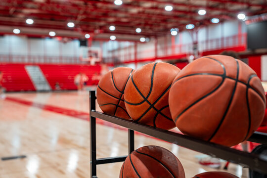 Close up photo of basketballs on storage rack inside of a gym with wood floors and red bleachers.
