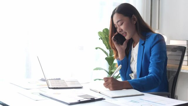 A woman in a blue suit is talking on her cell phone while writing in a notebook. She is focused on her work and is multitasking by handling both tasks at the same time