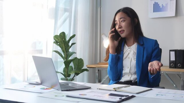 A woman in a blue suit is talking on her cell phone. She is looking at a laptop computer and has a pile of papers on her desk