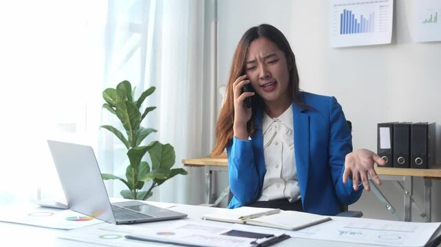 A woman in a blue suit is talking on her cell phone. She is sitting at a desk with a laptop and a stack of papers. The scene suggests a busy workday