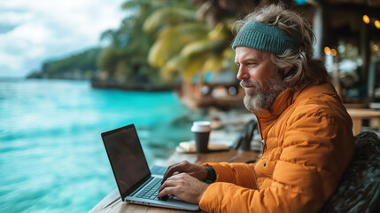 A bearded man in an orange jacket works on a laptop by a tropical waterfront, blending productivity with paradise.