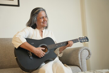 Happy mature grey hair indian woman singing and playing acoustic guitar at home. Retired Senior...