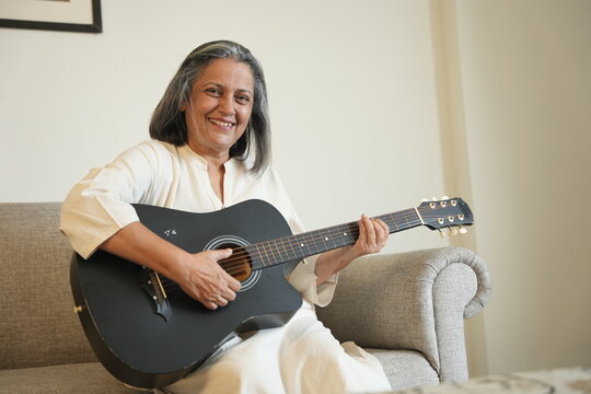 Happy mature grey hair indian woman singing and playing acoustic guitar at home. Retired Senior lady have fun with musical instrument wile sitting on sofa, Hobby and leisure activity. Mental health.