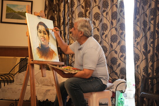 Indian creative senior artist man draw painting of woman on canvas with a brush and palette at home. Retirement life. Imagination, Creativity and art.