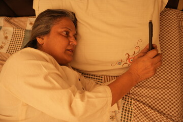  Senior indian woman using a smart phone while lying on bed late in night, Mature Addicted woman suffering from insomnia surfing on the internet with mobile phone, Social media obsession. 