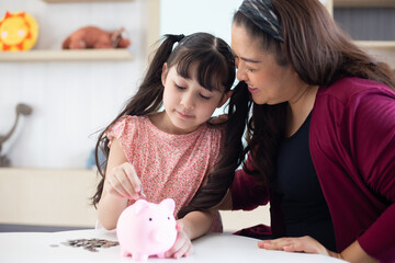 Smiling mother helping daughter putting money in piggy bank, .Cute little girl saving money by...