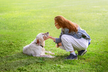 A woman squats in front of a white dog and says warm words to it against a background of green grass illuminated by sunlight. Close-up. Side view
