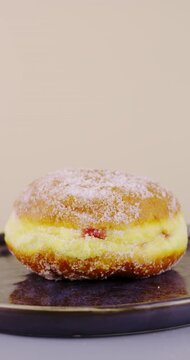A vertical video of a Berliner donut rotating on a dark plate. The donut is dusted with sugar, revealing its fluffy golden dough and a glimpse of red fruit filling, set against a light background.