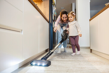 Small daughter with her mother is vacuuming and cleaning kitchen floor at home.