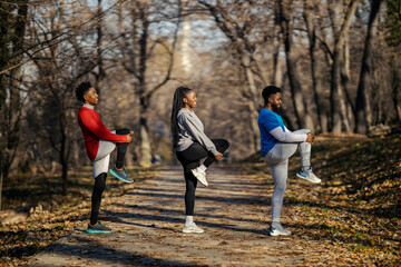 Group of three interracial sporty friends stretching legs and doing warmup exercises in nature.
