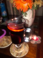 glass mug filled with a warm, dark liquid, possibly mulled wine or tea, garnished with an orange slice. The mug is placed on a wooden coaster on a table.